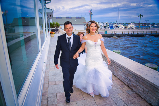Bride and groom walk hand-in-hand by the water.