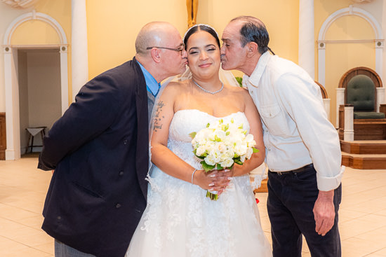 Bride kissed by two men in church setting.