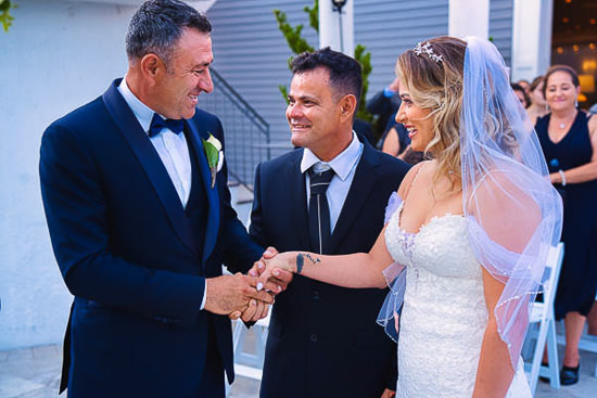 Bride shaking hands with father, groom smiling nearby.
