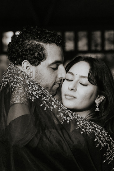 In a dimly lit room, a man and woman embrace. he gently nuzzles her cheek as she closes her eyes, seemingly lost in the moment. they are dressed in elegant, dark clothing with delicate embroidery.
