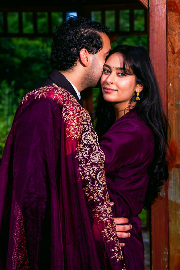 In a gazebo with a blurred green background, a man in a dark suit and a woman in a purple dress and shawl stand close together. the man kisses the woman's forehead, and she looks at the camera.