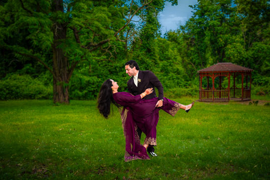 A grassy park with trees and a gazebo a man and woman dressed in formal south asian attire the man is dipping the woman