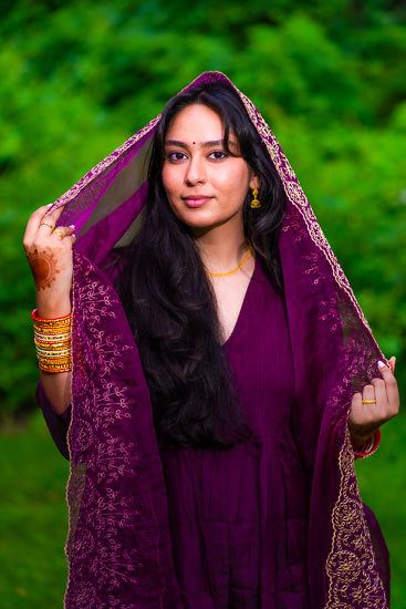 A young woman in traditional indian clothing holds her sheer purple dupatta over her head