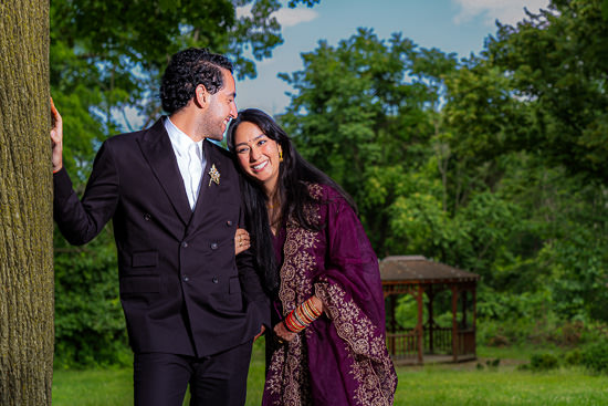 Outdoor park setting man and woman dressed for special occasion lean against tree he has arm around her they look at each other and smile